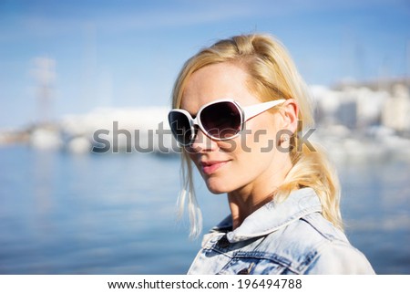 Beautiful natural young blond woman in sunglasses standing overlooking the ocean with the coastline behind her turning to smile at the camera, head and shoulders portrait