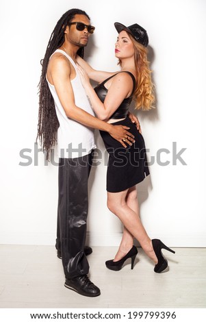 young multiracial couple with a dreadlock African American man wearing sunglasses and cute blond woman wearing dress against the wall kissing