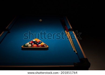 Pool balls stacked in the triangular rack, the cue ball and cues on a blue baize table in a shadowy dark nightclub waiting for the next players