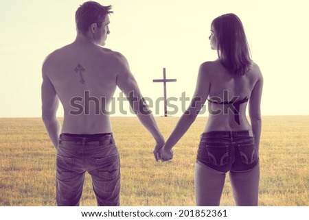 Atmospheric conceptual image of the silhouettes of a romantic young couple holding hands standing with their backs to the camera in a field with a cross visible beyond them