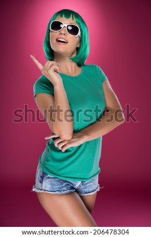 Trendy modern young woman with green hair wearing trendy sunglasses pointing up above her head with her finger looking up with a smile