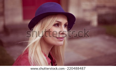 Young blond woman in a trendy hat standing looking off to the right of the frame with a quiet smile on her face