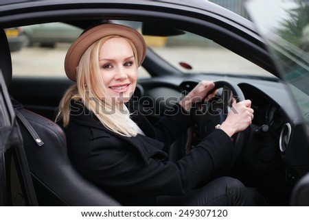 Attractive woman driver in a stylish hat sitting behind the steering wheel in her car looking through the open door at the camera with a friendly smile