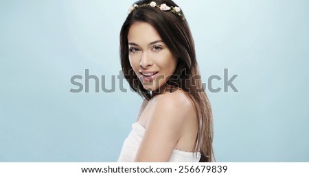 Pretty Smiling Young Woman with Small Flower Headband Wrapped in a Bath Towel While Looking at the Camera. Isolated on a Sky Blue Background