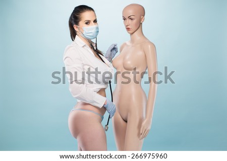 Sexy Female Nurse Wearing White Long Sleeves Shirt and Underwear with Mask, Gloves and Stethoscope Standing Besides a Human Dummy on a Very Light Blue Background.