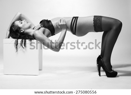 Slim brunette woman wearing black sexy underwear, lace high socks and high heel shoes, while posing in a provocative bridge position with head and shoulders on a cube, with copy space on gray