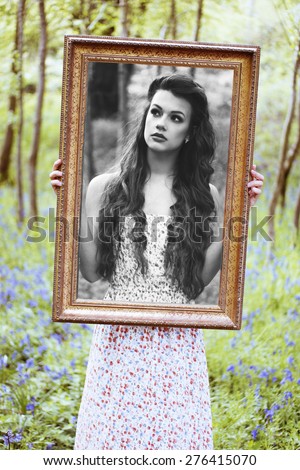 Ravishing beautiful redhead long haired woman wearing a low-cut sleeveless dress with floral pattern while looking at camera through a brown handheld portrait frame in a deciduous forest