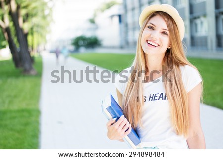 Cheerful Blond Student Teen Girl in Casual Outfit, Holding Books and Smiling at the Camera While Standing at the Outdoor Walkway.