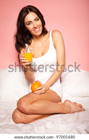 Smiling beautiful brunette woman curled up on her unmade bed with a glass of orange juice and a fresh orange for breakfast