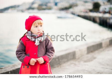 Portrait of little girl in the red coat and beret walking near sea in the city