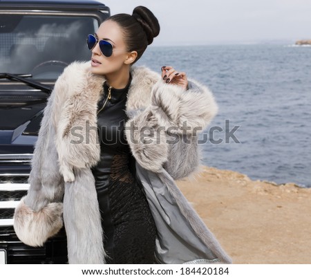 sexy glamour woman with dark hair posing beside a car in fur coat