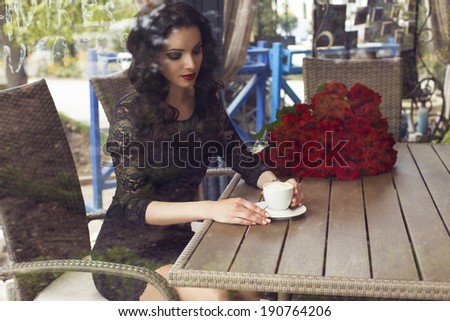 beautiful smiling woman with dark hair in black lace dress sitting in city cafe behind a glass window with cup of coffee and bouquet of red roses on background.
