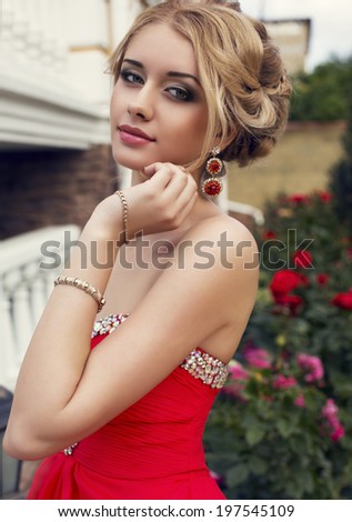 portrait of beautiful young woman with blond hair in elegant red dress posing at park