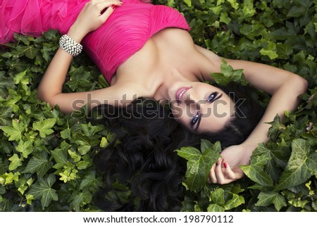 beautiful smiling woman in luxurious pink dress lying on green grass at summer garden