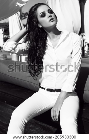 black and white fashion photo of sexy glamour model with long dark hair in elegant white clothes sitting on beach
