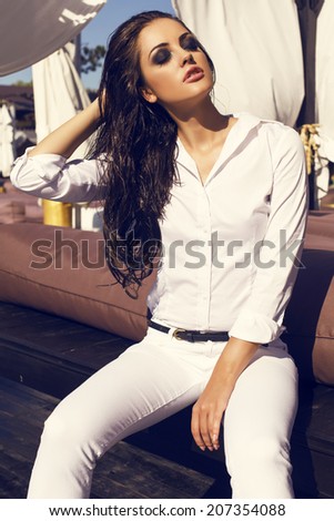 fashion photo of sexy glamour woman with long dark wet hair in white shirt and pants sitting on beach