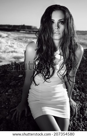black and white fashion photo of sexy beautiful woman with long dark wet hair in elegant white dress posing beside a sea