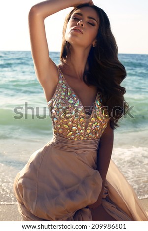 fashion photo of sensual beautiful woman with dark hair in luxurious sequin dress posing on summer beach in sunlight rays