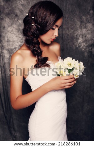 fashion photo of beautiful elegant bride with dark hair in elegant dress holding a wedding bouquet and posing at studio