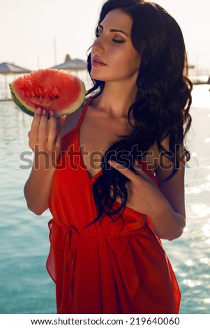 fashion outdoor photo of beautiful sensual woman with dark hair in elegant red dress eating watermelon beside a swimming pool