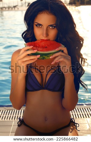 fashion outdoor photo of sensual beautiful woman with dark hair in blue swimsuit eating watermelon beside a swimming pool