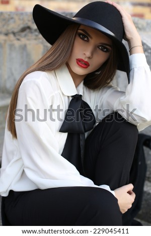 fashion outdoor photo of beautiful ladylike woman with dark straight hair wearing elegant blouse and felt hat, sitting on stair in autumn park