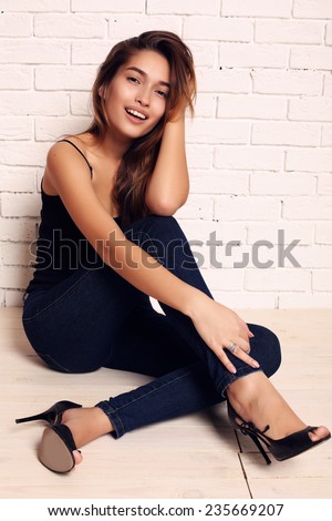 fashion indoor photo of beautiful smiling girl with dark hair wearing jeans and shoes,sitting beside a wall in studio