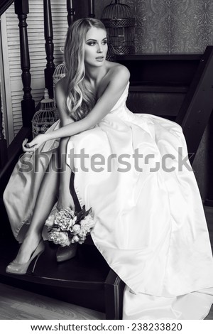 black and white fashion interior photo of beautiful bride with blond hair in elegant wedding dress,holding a bouquet of flowers,sitting on stairs
