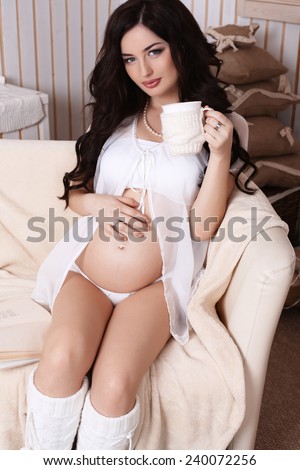 fashion photo of beautiful pregnant woman with long dark hair drinking cup of tea in cozy interior