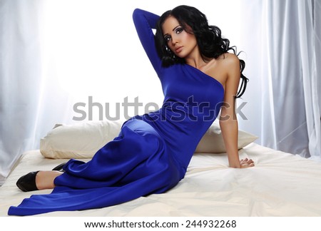 fashion photo of sexy glamour woman with dark hair wearing elegant blue dress, lying on bed in bedroom
