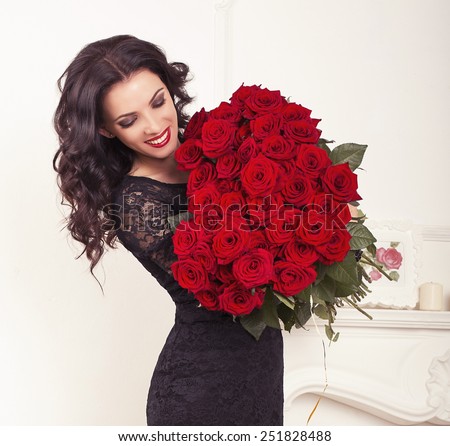 fashion interior photo of beautiful smiling woman with dark hair in elegant lace dress, holding a big bouquet of red roses in Valentine\'s day
