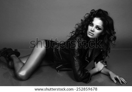 black and white fashion studio portrait of gorgeous young woman with dark curly hair wearing leather jacket and shoes