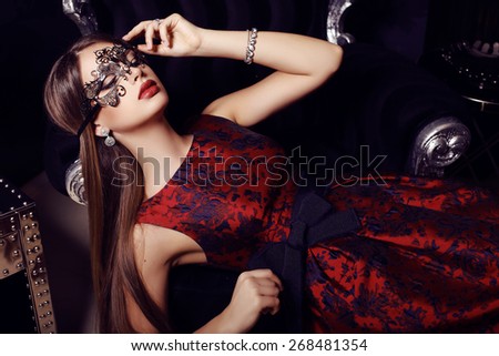 fashion photo of gorgeous woman with dark hair  in elegant dress and mask on face posing in luxurious interior