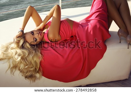 fashion outdoor photo of beautiful sexy woman with blond hair wearing elegant pink dress , relaxing on yacht