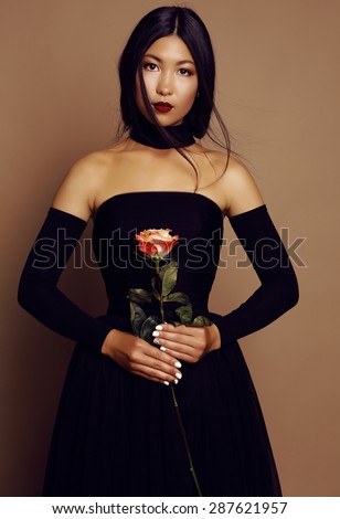 fashion studio photo of beautiful asian look woman with dark hair wearing elegant black dress,holding one rose in hands
