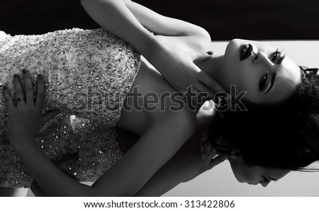 black and white fashion outdoor photo of beautiful young woman with dark hair in luxurious sequin dress lying on mirror