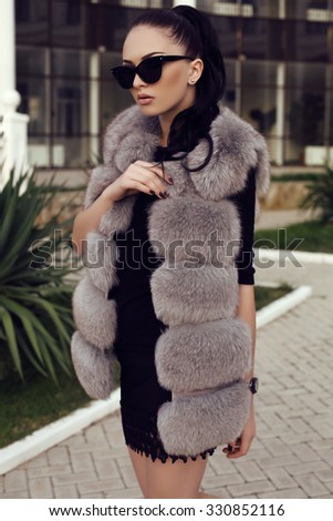 fashion outdoor photo of gorgeous woman with long dark hair wears luxurious fur coat and sunglasses, walking by the street