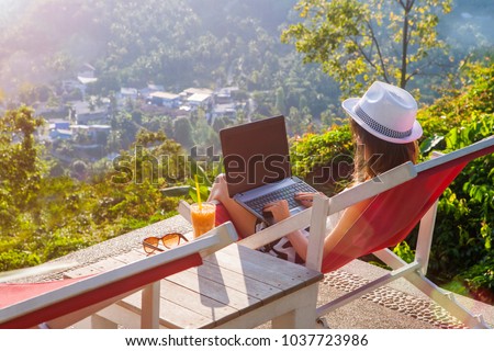 Girl freelancer work typing on laptop and looking at monitor with beautiful view and cocktail outdoors sun sky and greens. Traveling with a computer. Online dream job on the island in Thailand concept