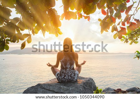 Woman back side meditating sitting on big rock doing yoga in lotus pose under a tree and looking at the sun rays of the sunset, bright rays enlightenment, opening chakras bright colorful concept
