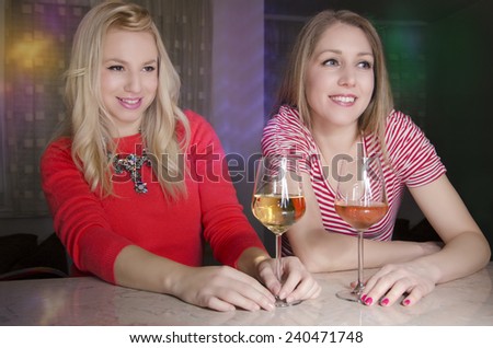 Two beautiful girls sitting by the bar and drinking wine, looking at somebody and smiling, flirting, selective focus on second girl