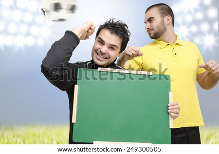 Man with empty blackboard cheering for scored goal, fist in the air, soccer player in action in background, selective focus