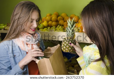 Two beautiful, natural looking girls buying fruit in greengrocery, holding avocado and pineapple