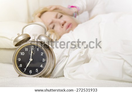 Blonde girl holding hands on her ears, because of alarm clock sound. Selective focus on clock
