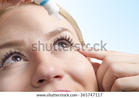 Closeup of young woman applying eye drops, selective focus only on right eye. Drop captured in mid air/ eye drops with vitamins/ eye care