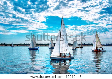 Start of a sailing regatta, fully crewed yachts with white sails catching the wind, blue sky and white clouds on background.