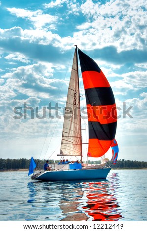 Start of a sailing regatta, fully crewed yacht with black and red sails catching the wind, blue sky and white clouds on background.
