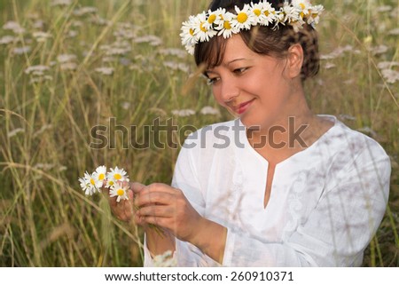 Smiling young woman makes daisy diadem on meadow