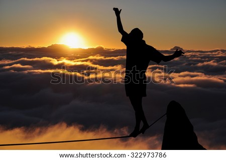 Silhouette of young man balancing on slackline high above clouds during sunset. Slackliner balancing on tightrope, highline silhouette.