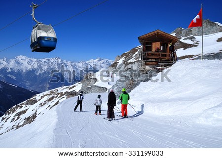 Skiers on the ski slope in Swiss Alps in sunny day. Small wooden house with red swiss flag near by. Blue cable car above and mountains behind, skiing resort Thyon 2000, Switzerland.