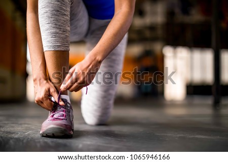 Woman tying her shoe in an open empty gym, preparing for workout or run, wearing gray leggings and royal blue top.